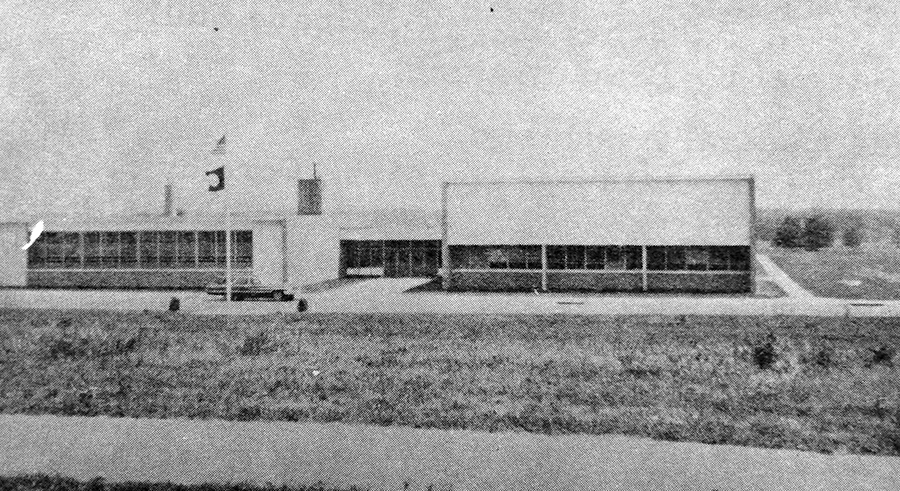 Black and white photograph of the main entrance to Hayfield Elementary School taken in 1967. The multi-purpose room and cafeteria are on the left, and the classroom wing and offices are on the right. A station wagon is parked in front of the building. A sidewalk runs down the side of the building where an addition will be constructed in 1972. Two small trees or shrubs have been planted near the flagpole.  