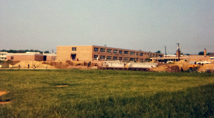 Color photograph of the exterior of Hayfield Elementary School taken from the playing fields behind the school. Construction work is in progress because there are large mounds of earth between the field and the school. The open classroom wing is complete, but the gymnasium and music room addition are not yet constructed. 
