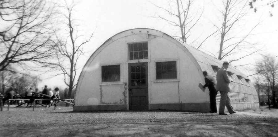 Black and white photograph taken in 1954 of a Quonset hut at Fairfax Elementary School. The building is a long semi-cylindrical structure with a door and three windows on one end and four windows along each side. The buildings were made out of metal and could be rapidly assembled and disassembled. Children can be seen to the left of the Quonset hut playing on the playground. Two men are examining the structure.  