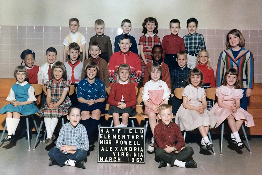 Color class photograph from the 1966 to 1967 school year. A sign indicates the teacher’s name was Miss Powell and that the picture was taken in March 1967. 23 children are pictured, an even mix of boys and girls. Some are standing, others are seated. The girls are all wearing dresses or skirts and the boys are wearing button down shirts or sweaters. Miss Powell can be seen standing on the right. She has shoulder length blonde hair and is wearing a multi-colored vertical striped blazer and crew neck blue blouse. 