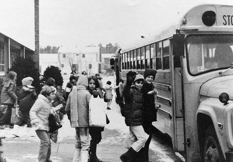 Black and white yearbook photograph taken in 1980 showing students at dismissal. It is wintertime and there is snow on the ground. The children are wearing heavy coats and are boarding school buses. One of the children is waving at the camera. 