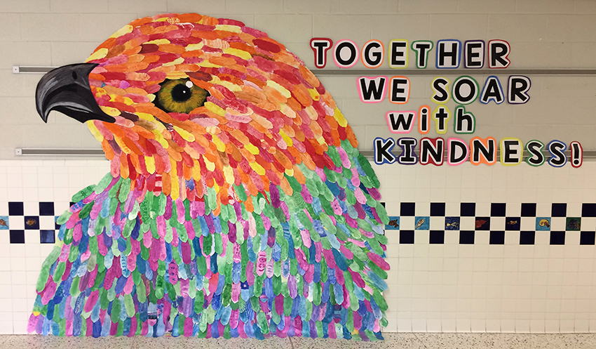 Photograph of artwork created by students in 2018 displayed in a hallway at Hayfield Elementary School. Created to look like a hawk, this large piece of artwork is approximately ten feet wide and covers the wall from floor to ceiling. Pieces of yellow, orange, red, green, blue, and lavender paper, cut into long oval shapes, have been overlapped to look like feathers. Next to the hawk, text printed in large letters reads: Together We Soar with Kindness! 