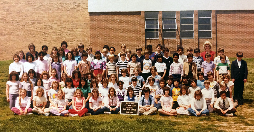 Color photograph of Hayfield’s sixth grade class. They are posed outside of the building with some children seated on the ground and others standing behind them. In the background several teachers can be seen standing behind the students. Approximately 80 children are pictured.